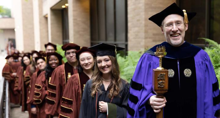 People wearing graduation cloaks and smiling. One man is holding a graduation mace.