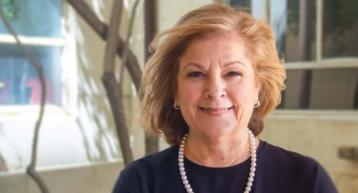 Diane Ginsburg wearing pearls and smiling while standing in front of a tree and building.