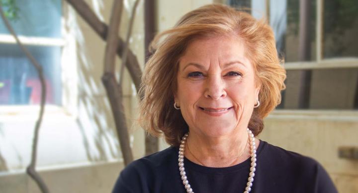 Diane Ginsburg wearing pearls and smiling while standing in front of a tree and building.