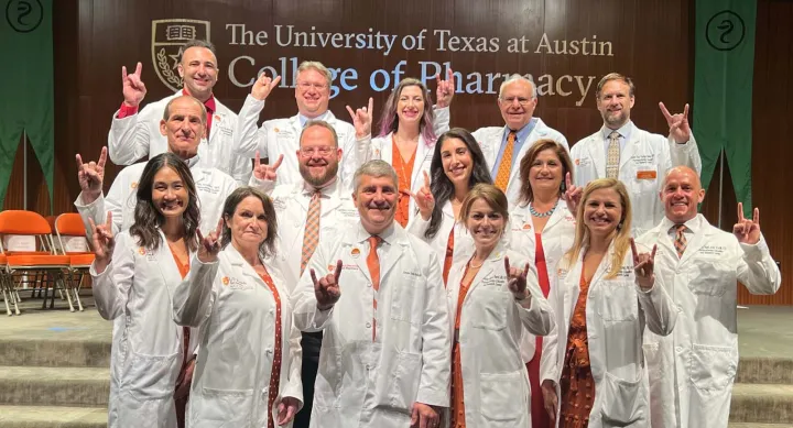 A group of people posing in white coats and giving the Hook 'em Horns hand gesture in front of the UT Austin College of Pharmacy wordmark.