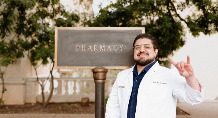A headshot of student pharmacist Erik Coronado. Erik poses outside the Pharmacy building by the Pharmacy sign. He has brown hair and a brown beard and glasses. Erik holds his hands with "horns up" and smiles to camera in his white coat.