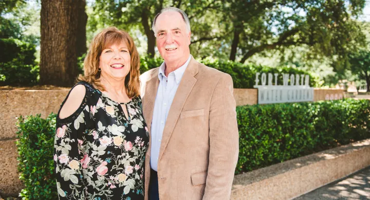 Dr. Polansky and his wife Barbara pose outside against a green shrub. Dr. Polansky wears a tan suit jacket and Barbara a black and floral patterened shirt. They are both smiling to camera.
