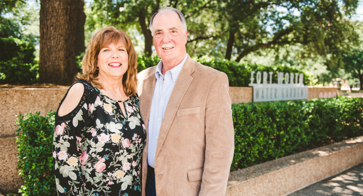 Dr. Polansky and his wife Barbara pose outside against a green shrub. Dr. Polansky wears a tan suit jacket and Barbara a black and floral patterened shirt. They are both smiling to camera.