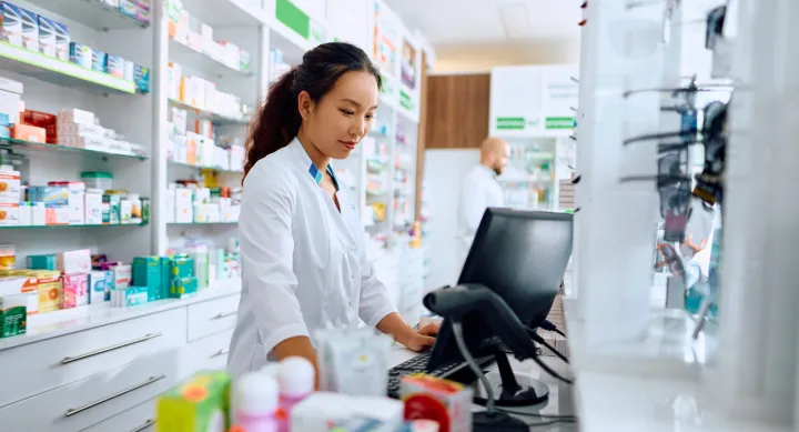 A stock image of a female pharmacist working at the Pharmacy counter. She has a brown pony tail and is wearing a white coat.