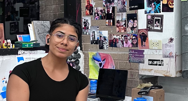 Student artist Pierce Cedillo sits in her studio and smiles to camera. She is wearing glasses and a black tshirt, with her dark hair pulled back into a bun. Behind her is her "Inspiration Wall" - a backdrop of photos, doodles, and ideas.