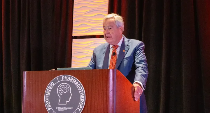 Dr. M. Lynn Crismon stands on stage at the podium giving a presentation. He's wearing a grey suit with a burnt orange tie.