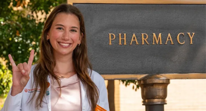 A woman standing in front of a pharmacy sign and giving the Hook 'em Horns hand gesture.