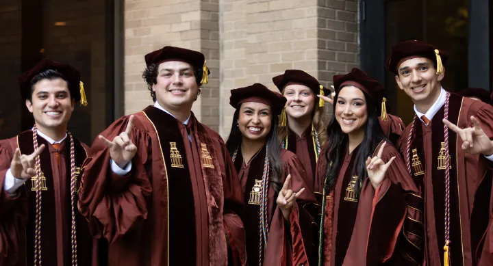 A group of graduating students in their regalia giving the Hook 'em Horns.