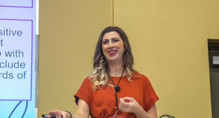 Dr. Sarah Kubes gives her presentation at the Preceptor Conference. She's wearing a short-sleeve burnt orange shirt and is smiling at the crowd. 