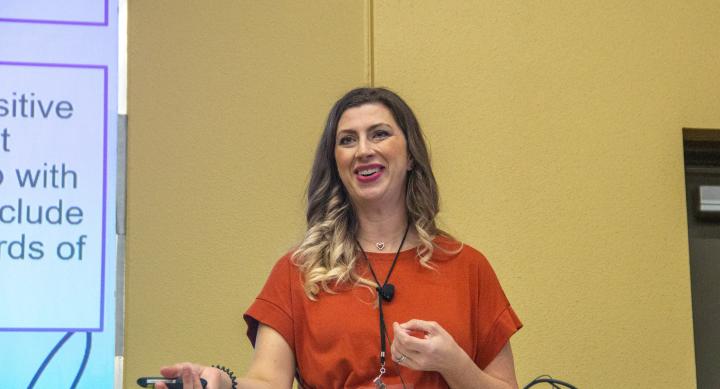 Dr. Sarah Kubes gives her presentation at the Preceptor Conference. She's wearing a short-sleeve burnt orange shirt and is smiling at the crowd. 
