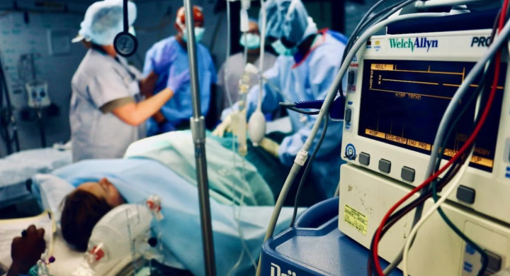A patient lies on a table in a busy operating room filled with machinery while doctors, nurses and medical staff attend to the patient.