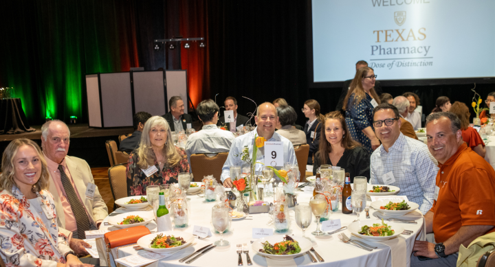 A group photo of Texas Pharmacy alumni, faculty and friends sitting together at a round gala table during a college event. All are smiling brightly to camera.