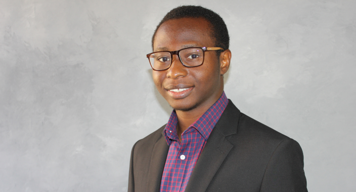 A headshot of graduate student Ayobami. He is smiling brightly to camera in a dark suit, purple shirt and rectangular glasses.