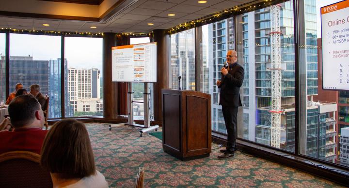 Dr. Glen Baumgart speaking at a lectern in front of windows with views of Austin skyscrapers