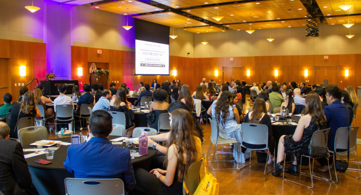 A banquet hall filled with attendees sitting at round tables facing a speaker at a podium. A large presentation slide reads "Celebration of Excellence. College of Pharmacy"